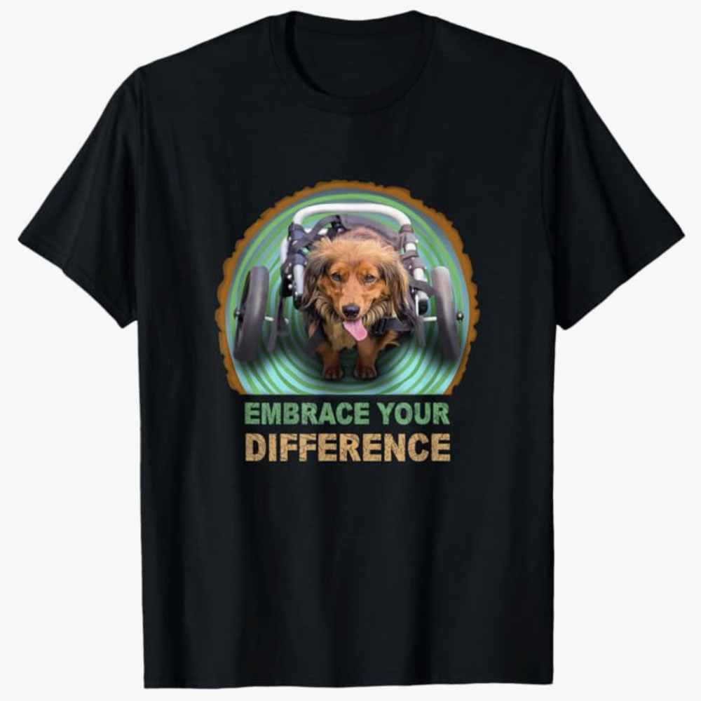 Black T-shirt featuring a happy dachshund using a wheelchair with the text Embrace Your Difference, promoting positivity, inclusion, and acceptance of disabilities through an inspiring and heartwarming design.