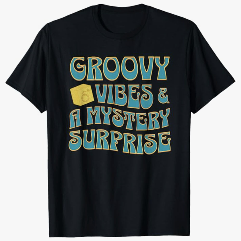 A black T-shirt features retro-style lettering reading Groovy Vibes & a Mystery Surprise with a small cube icon. The design conveys a fun, nostalgic mood paired with the excitement of an unexpected reveal.