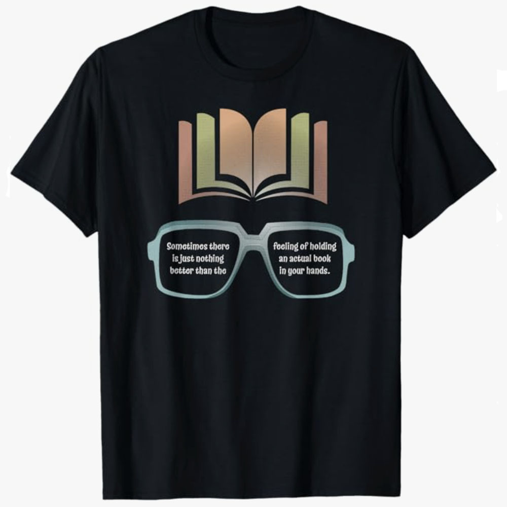 A black T-shirt features an open book above a pair of glasses with a message celebrating the joy of holding a real book. The design expresses a love for reading and the simple pleasure of physical books.