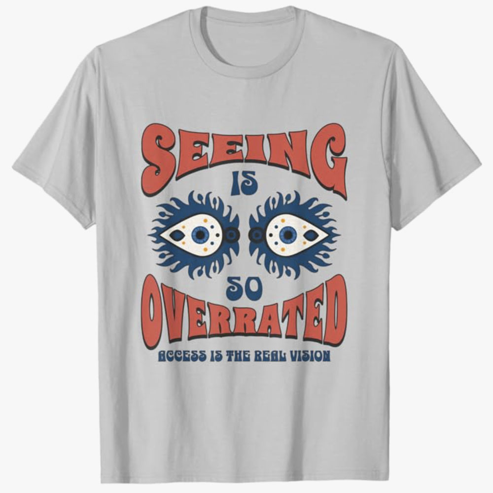 A light gray T-shirt reads Seeing Is So Overrated with illustrated eyes and the phrase Access Is the Real Vision, promoting disability awareness and the importance of accessibility.